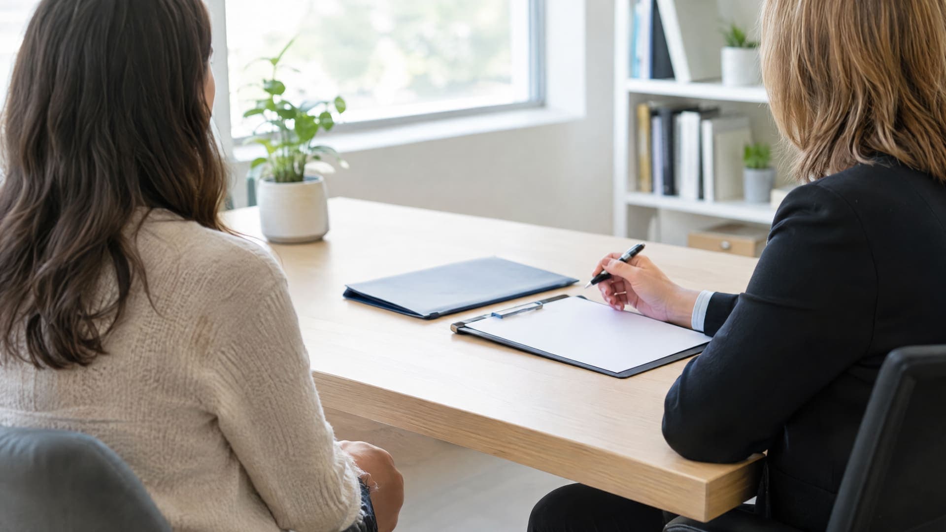 Beratungsgespräch an einem Bürotisch, zwei Personen von der Seite und von hinten, ein leeres Formular und eine Mappe auf dem Tisch