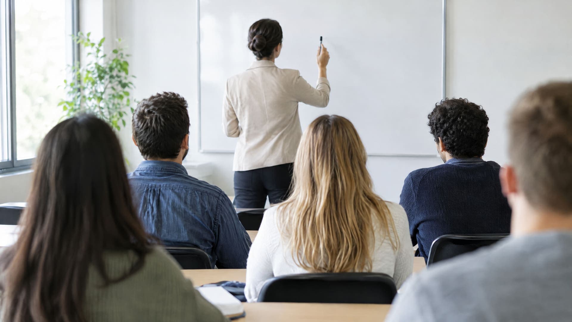 Dozent steht von hinten vor einer Gruppe erwachsener Lernender am Whiteboard in einem hellen Klassenraum