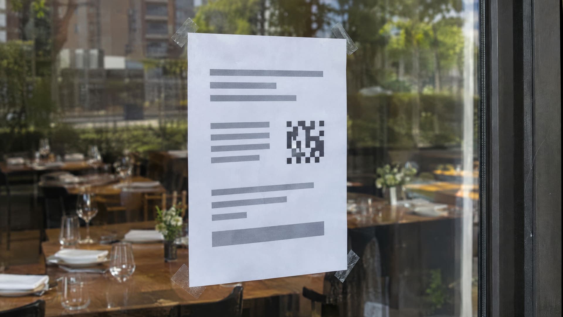 Zettel mit abstrakten grauen Zeilen und einem kleinen schwarzweißen Muster am Fenster eines Restaurants, dahinter unscharf gedeckte Tische im Tageslicht, keine Personen