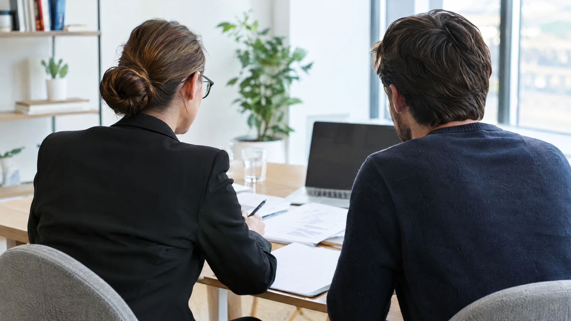 Zwei Personen von hinten sitzen an einem Tisch mit Unterlagen im Beratungsgespräch, eine Beraterin und ein Gründer, helles Büro, Gesichter nicht sichtbar
