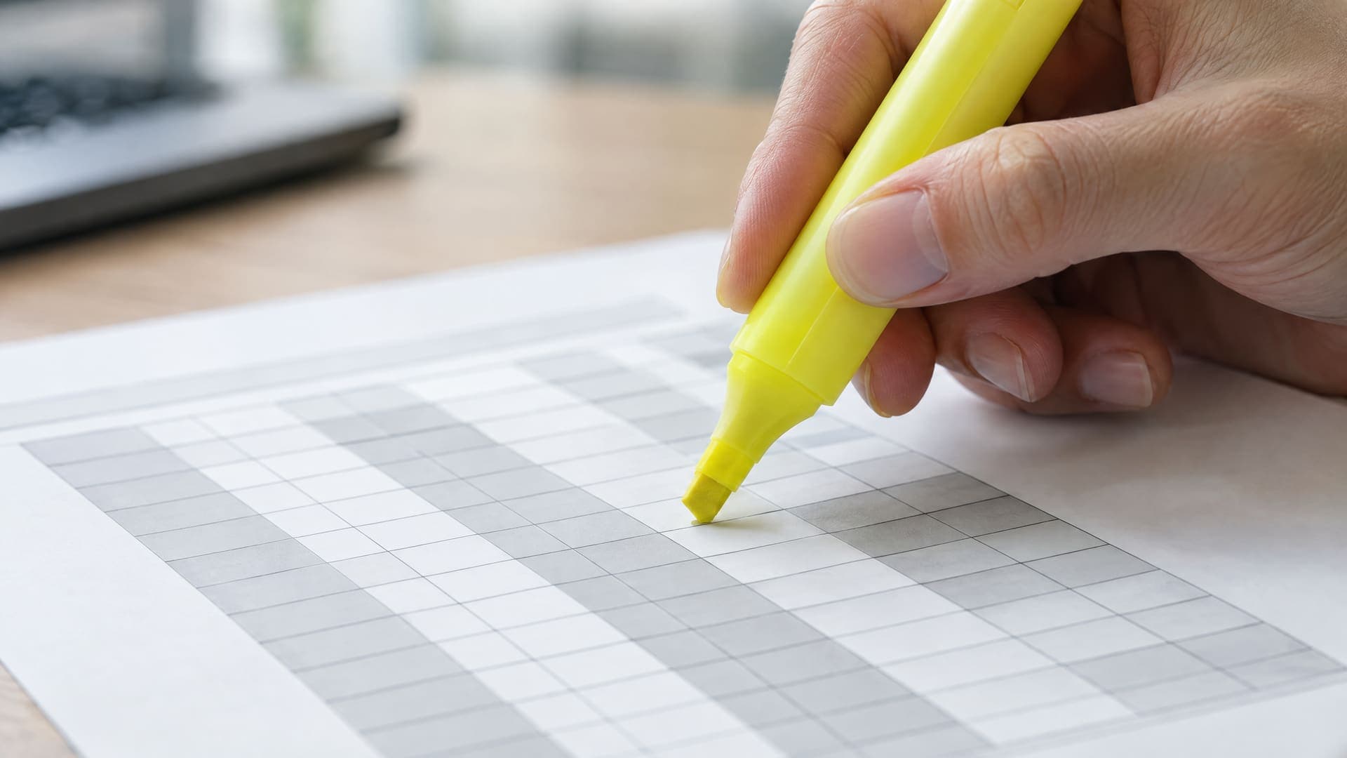 Hand mit gelbem Textmarker über einem ausgedruckten Blatt mit einem grauen Tabellenraster, Beschriftungen unleserlich