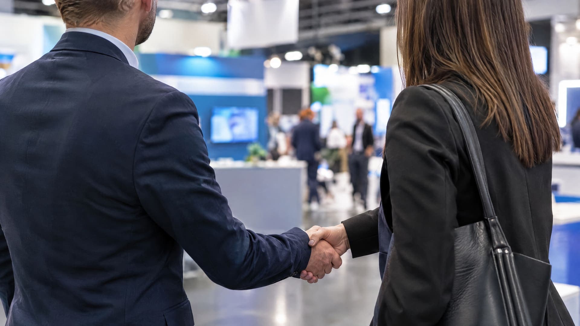 Handschlag zwischen zwei Geschäftsleuten an einem Messestand, von hinten fotografiert, unscharfe Messestände im Hintergrund, Gesichter nicht sichtbar