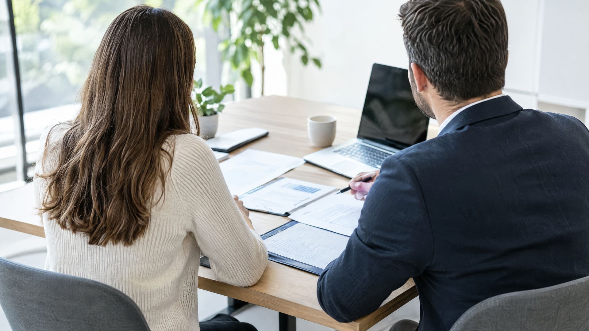 Zwei Personen von hinten an einem Tisch im Erstgespräch mit Unterlagen und einem Laptop in einem hellen Büro, Gesichter nicht sichtbar