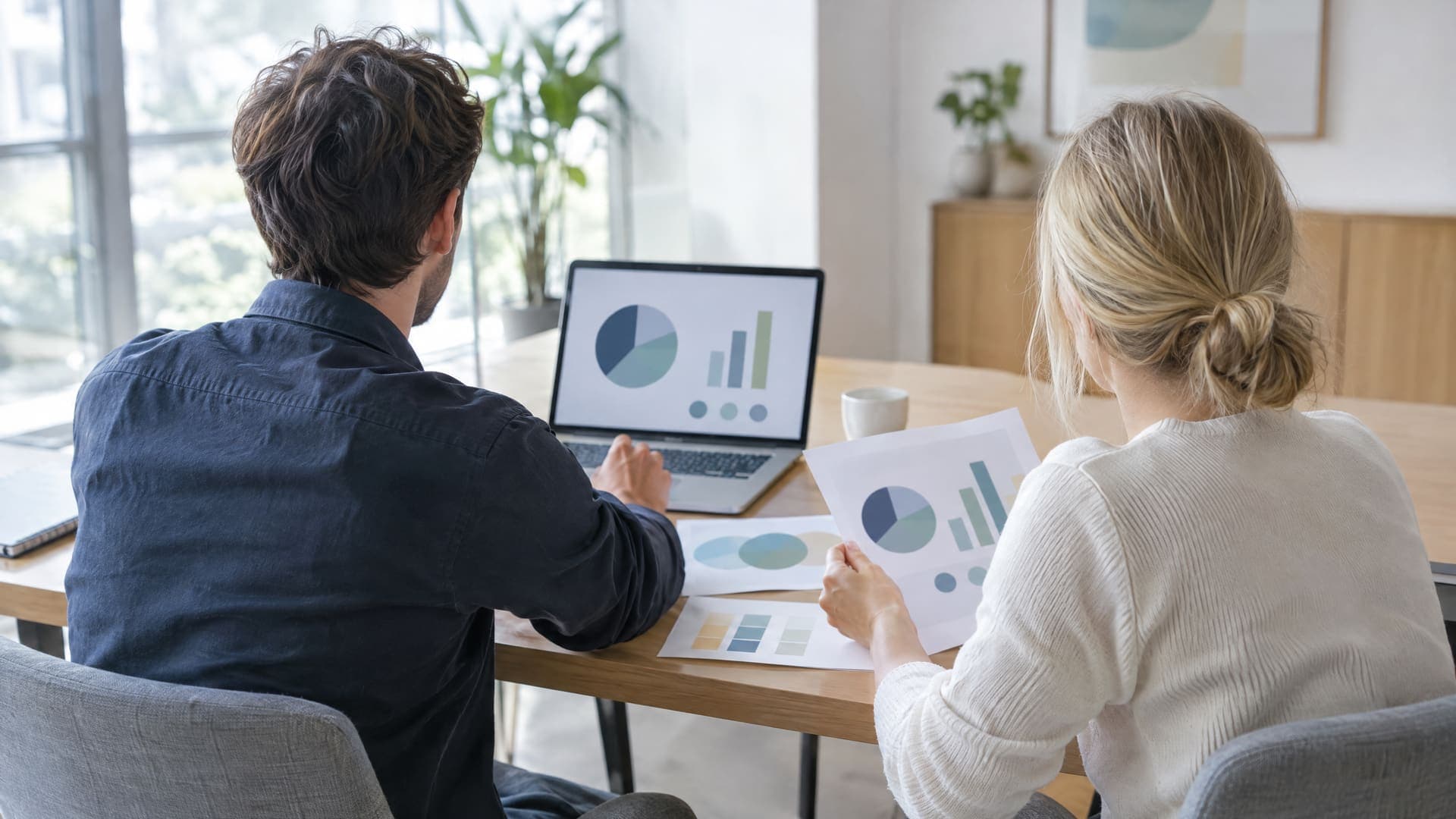 Zwei Personen von hinten an einem Tisch mit Laptop und Ausdrucken in einem Besprechungsraum, Gesichter nicht sichtbar