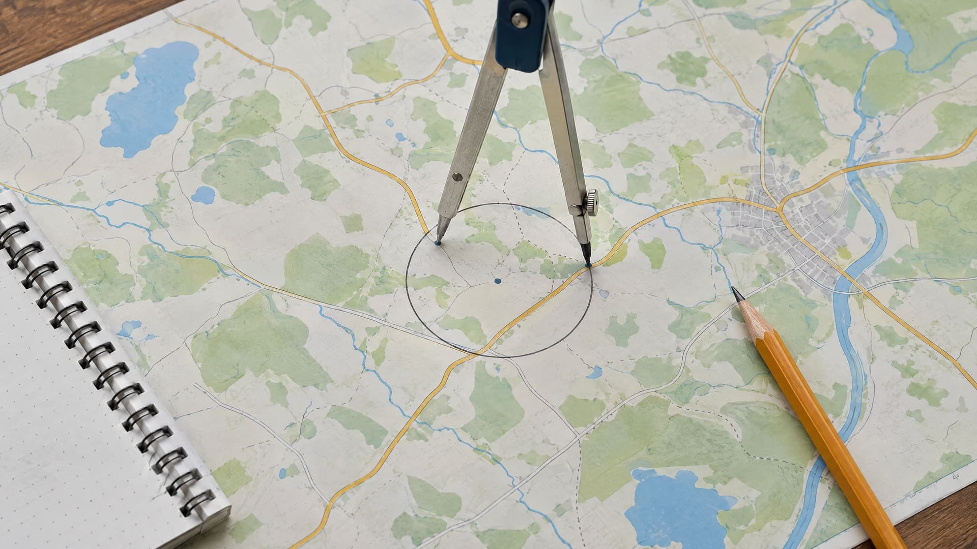 Zirkel zeichnet einen Kreis um einen Punkt auf einer Regionalkarte, daneben ein Bleistift und ein Notizbuch, keine lesbare Beschriftung