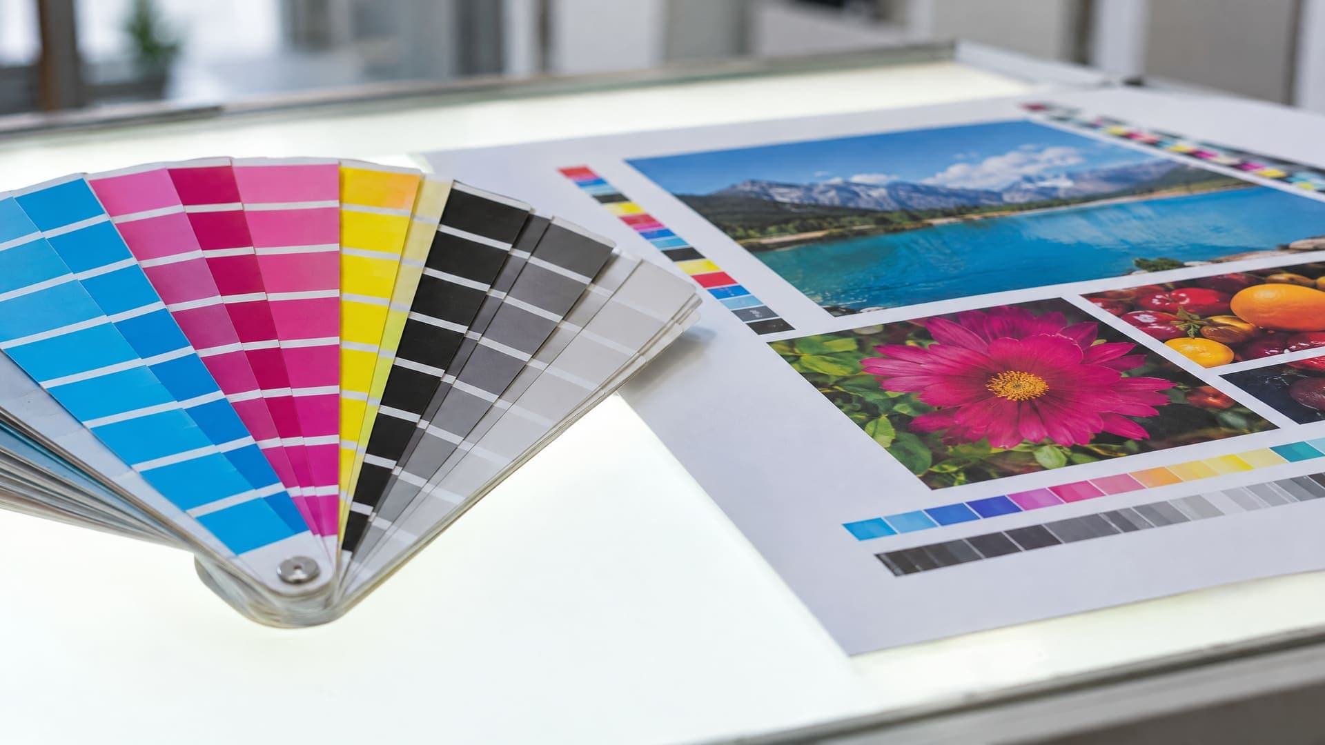 Fächer aus Cyan-, Magenta-, Gelb- und Schwarz-Farbmustern neben einem gedruckten Andruckbogen auf einem hellen Tisch im Tageslicht