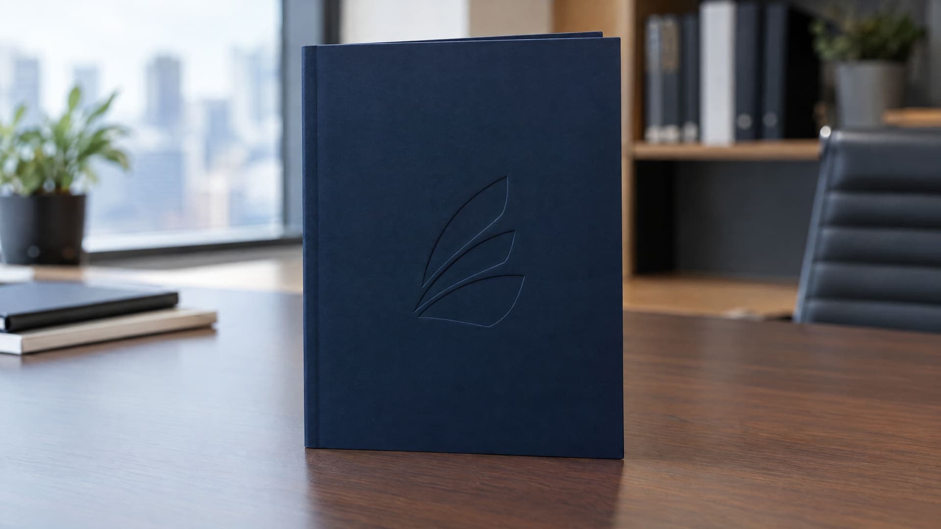 Broschüre aufrecht auf einem Schreibtisch, Umschlag in Navy mit Softtouch-Oberfläche und einem einzigen geprägten abstrakten Zeichen, Büro unscharf im Hintergrund