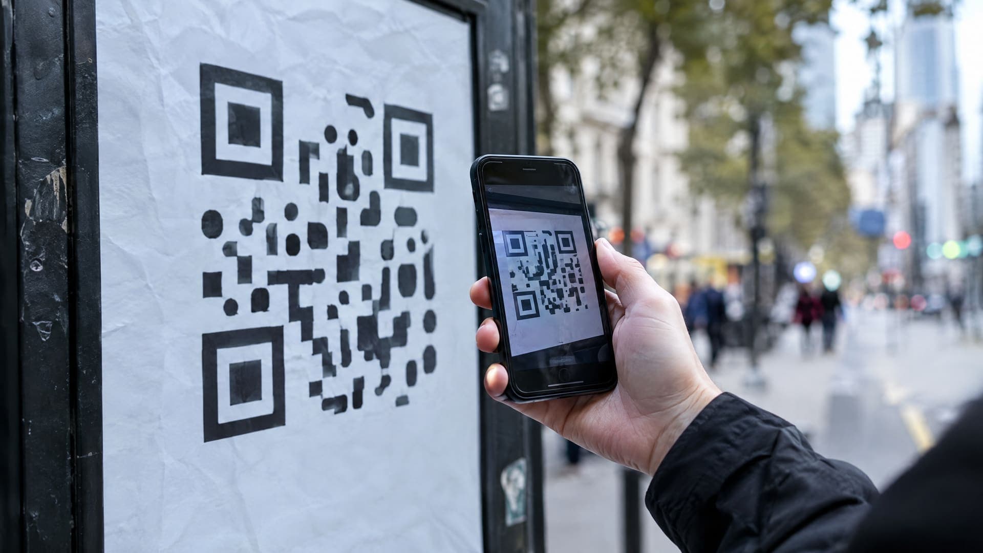 Hand hält ein Smartphone auf ein Straßenplakat mit abstraktem QR-Code, Straße unscharf im Hintergrund, Gesicht nicht sichtbar
