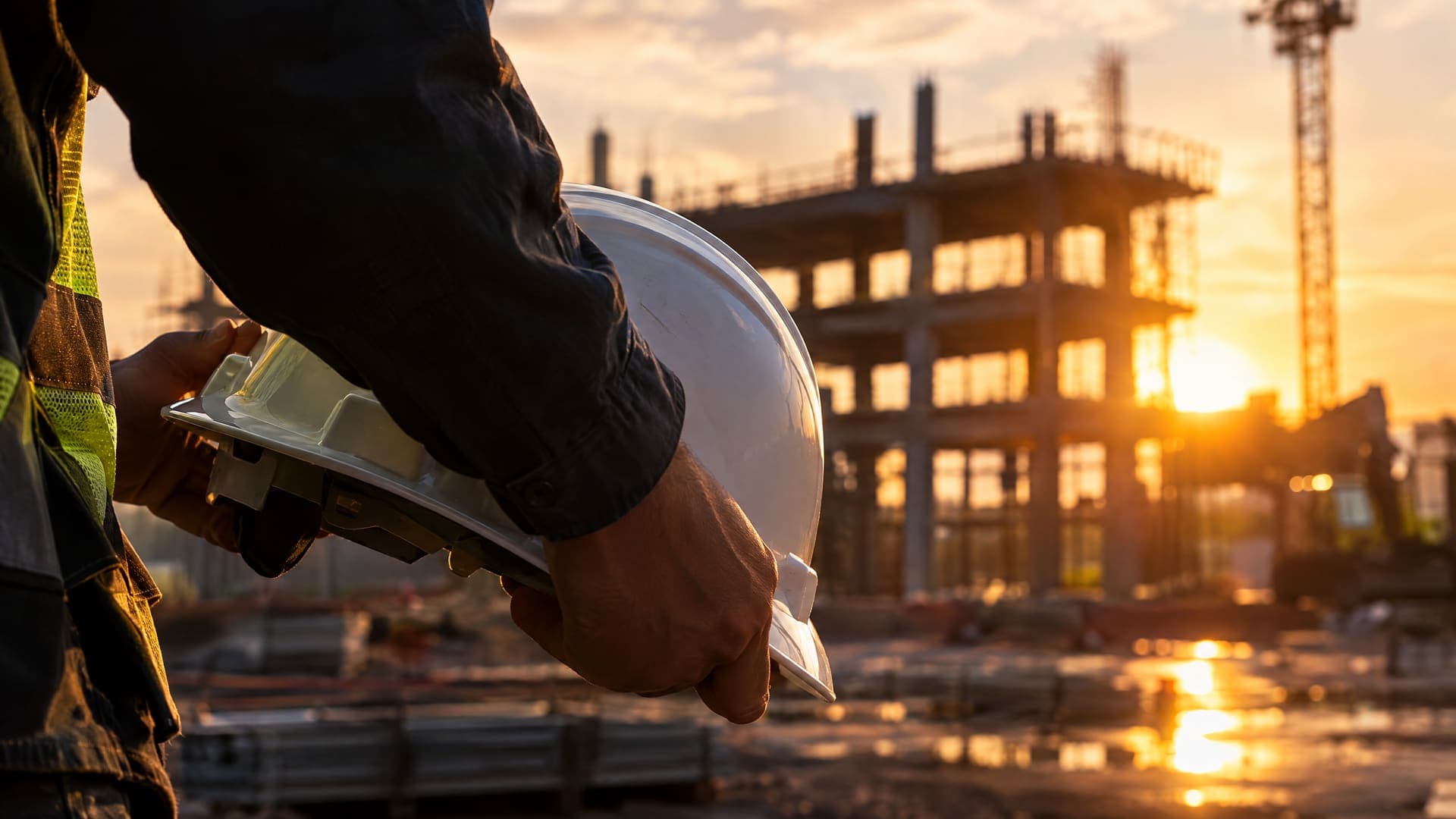 Hände halten einen Schutzhelm auf einer Baustelle im Gegenlicht, im Hintergrund unscharf ein Rohbau, keine Gesichter