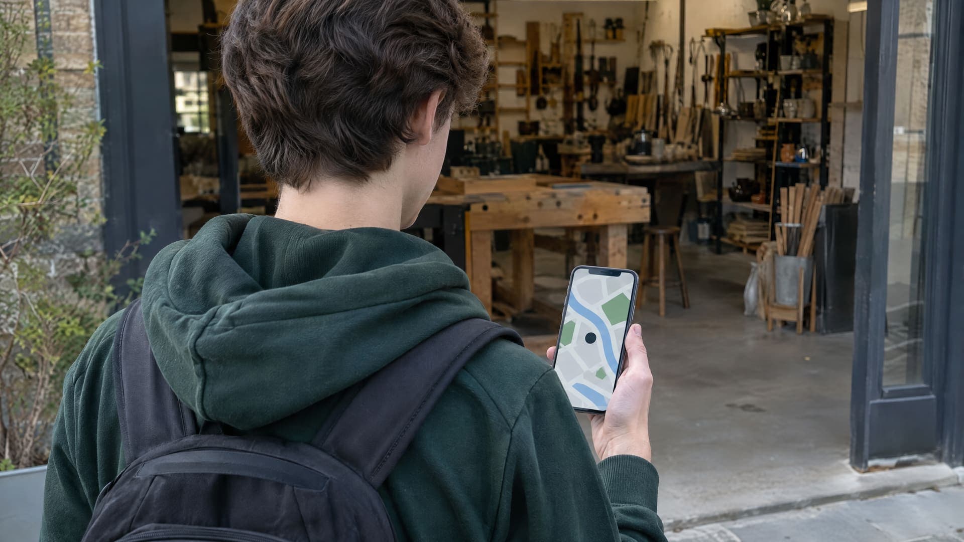 Jugendlicher von hinten steht mit Smartphone in der Hand vor einer Handwerkswerkstatt, auf dem Display eine abstrakte Kartenansicht, Gesicht nicht sichtbar