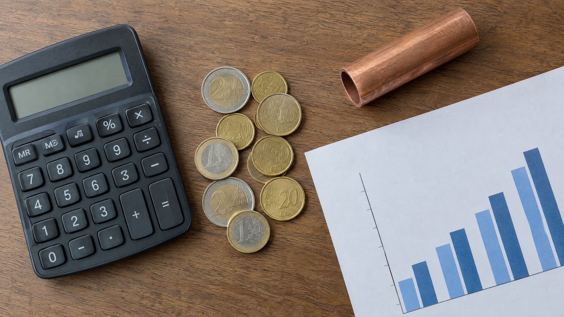 Draufsicht auf einen Taschenrechner, Euro-Münzen, ein kurzes Kupferrohrstück und ein Blatt mit Balkendiagramm auf einem Tisch