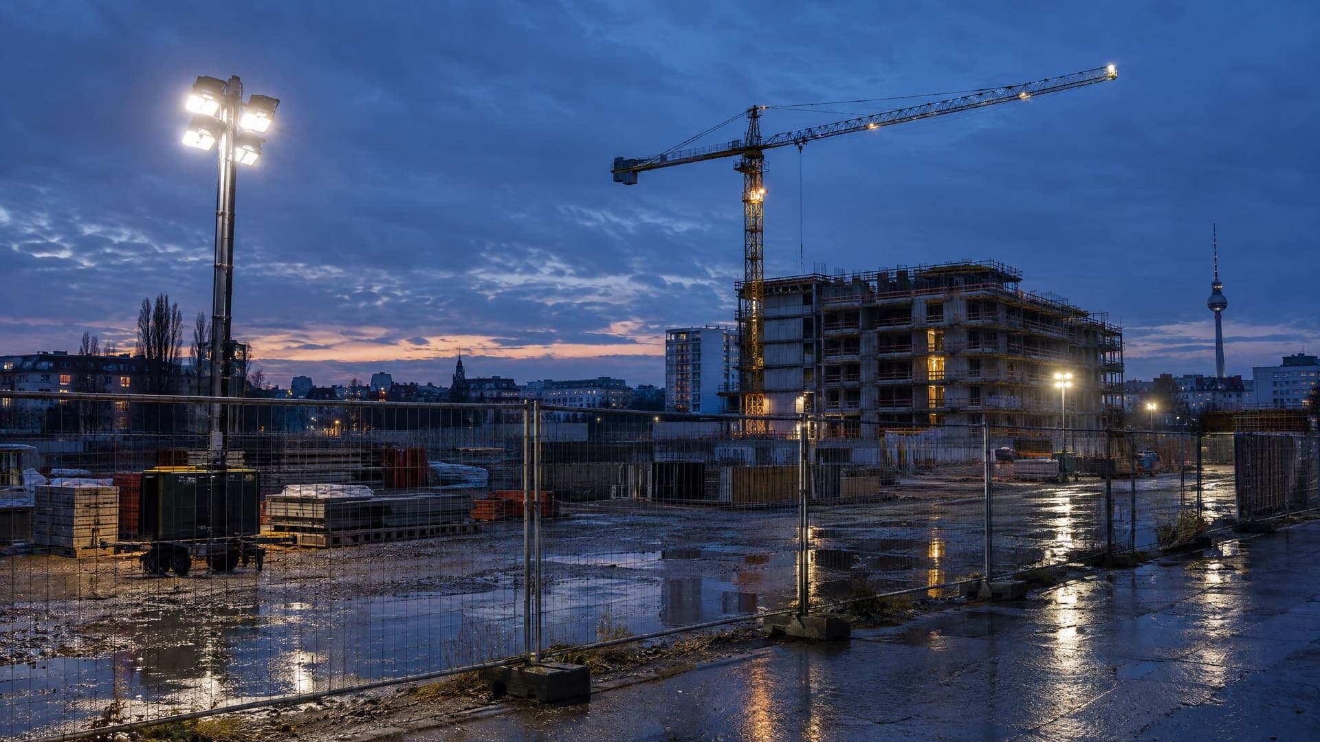 Bauzaun mit Flutlicht und Baukran vor einem Rohbau in Berlin in der blauen Stunde