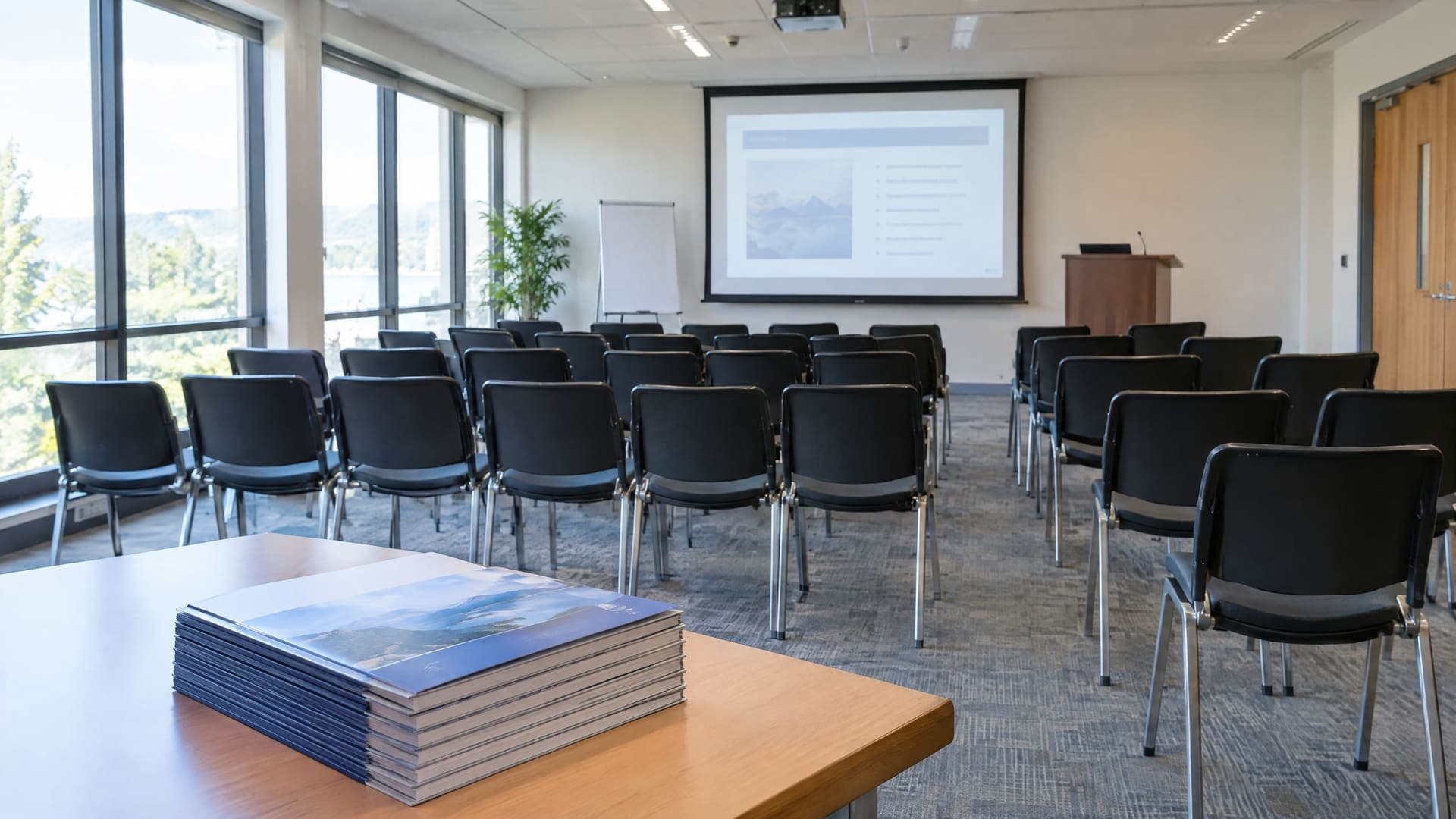 Leerer Schulungsraum mit Stuhlreihen, Präsentationsleinwand, Rednerpult und einem Stapel Arbeitsmappen auf einem Tisch