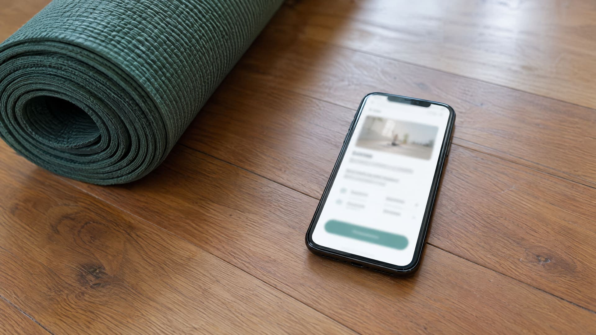 Smartphone mit einer Buchungsseite mit grünem Button auf einem Holzboden, daneben eine gerollte grüne Yogamatte