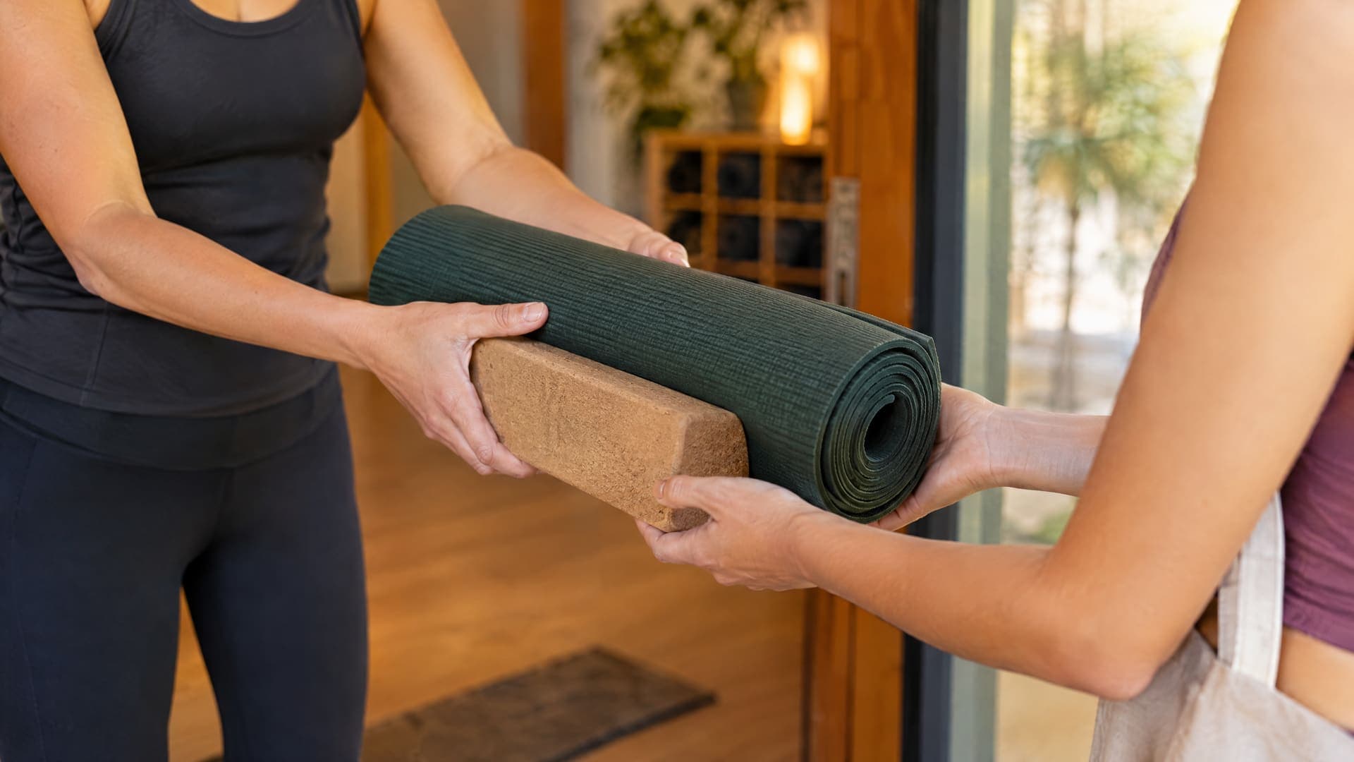 Zwei Personen am Eingang eines Yogastudios, eine übergibt einer anderen eine gerollte grüne Matte und einen Korkblock, nur Oberkörper und Hände sichtbar