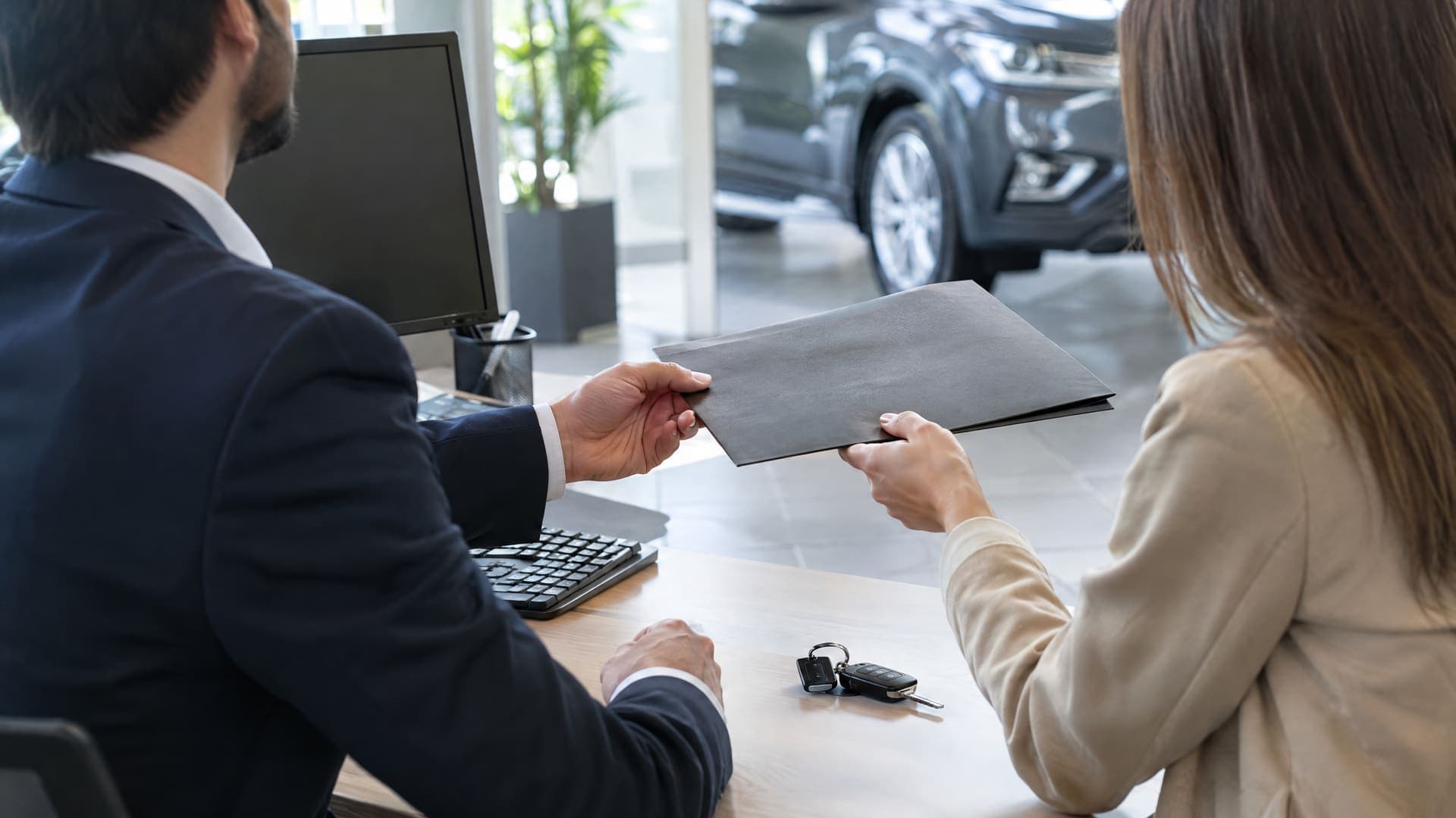 Zwei Geschäftsleute an einem Schreibtisch in einem Autohaus-Büro tauschen eine Mappe aus, von hinten und von der Seite fotografiert, Autoschlüssel auf dem Tisch